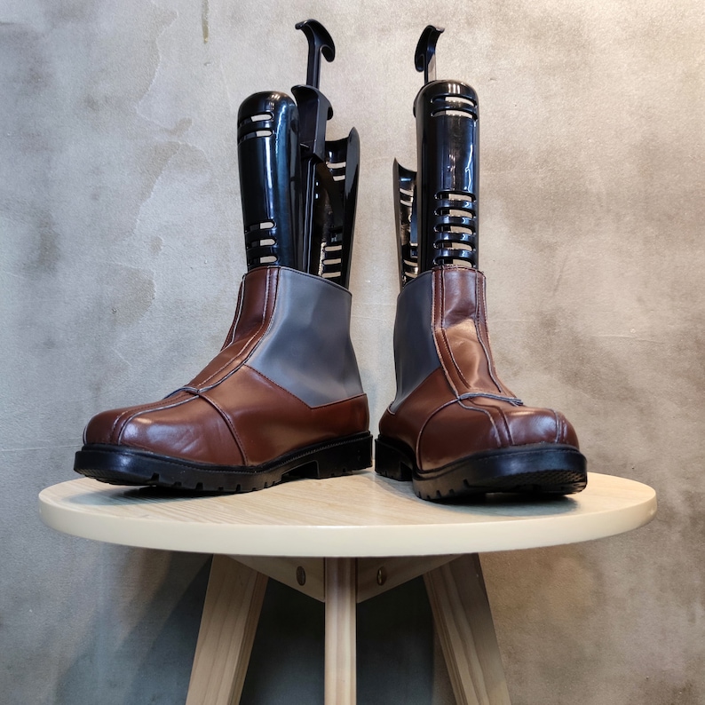 May include: A pair of brown leather boots with gray fabric tops. The boots are on a wooden table and have black boot shapers inside.