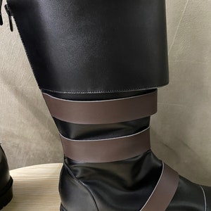 K&ouml;nnte beinhalten: Schwarze Lederstiefel mit braunen Riemen. Die Stiefel haben einen Rei&szlig;verschluss an der Seite und einen umklappbaren Schaft.