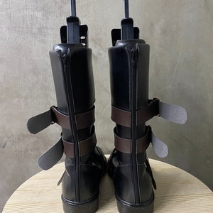 K&ouml;nnte beinhalten: Ein Paar schwarze Lederstiefel mit braunen Riemen und Schnallen. Die Stiefel haben einen Rei&szlig;verschluss an der R&uuml;ckseite und eine abgerundete Spitze.