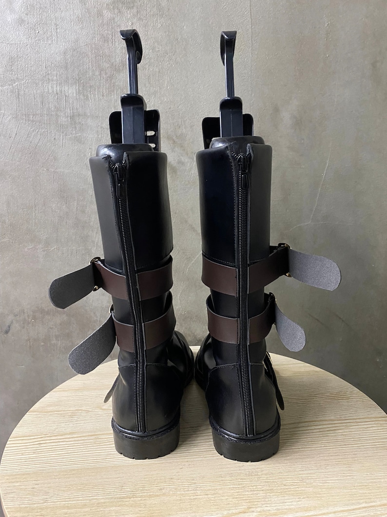 K&ouml;nnte beinhalten: Ein Paar schwarze Lederstiefel mit braunen Riemen und Schnallen. Die Stiefel haben einen Rei&szlig;verschluss an der R&uuml;ckseite und eine abgerundete Spitze.