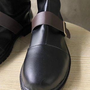 K&ouml;nnte beinhalten: Nahaufnahme eines Paares schwarzer Lederstiefel mit braunen Lederriemen und goldenen Schnallen. Die Stiefel haben eine abgerundete Spitze und eine dicke Sohle.