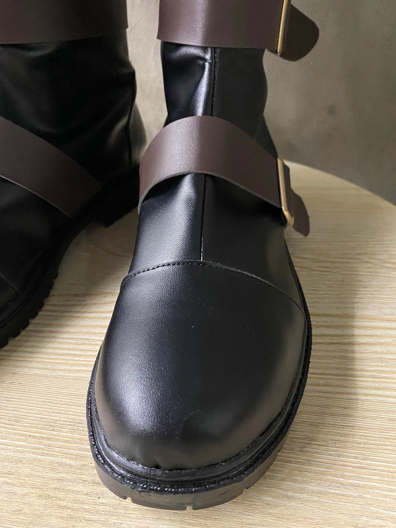 K&ouml;nnte beinhalten: Nahaufnahme eines Paares schwarzer Lederstiefel mit braunen Lederriemen und goldenen Schnallen. Die Stiefel haben eine abgerundete Spitze und eine dicke Sohle.