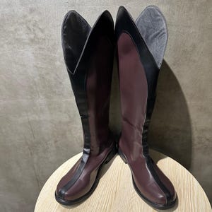 May include: Knee-high boots in burgundy and black. The boots have a polished finish, with a black stripe down the front and a flared top. The boots are displayed on a light-coloured, round surface. The boots are approximately 40 cm high.