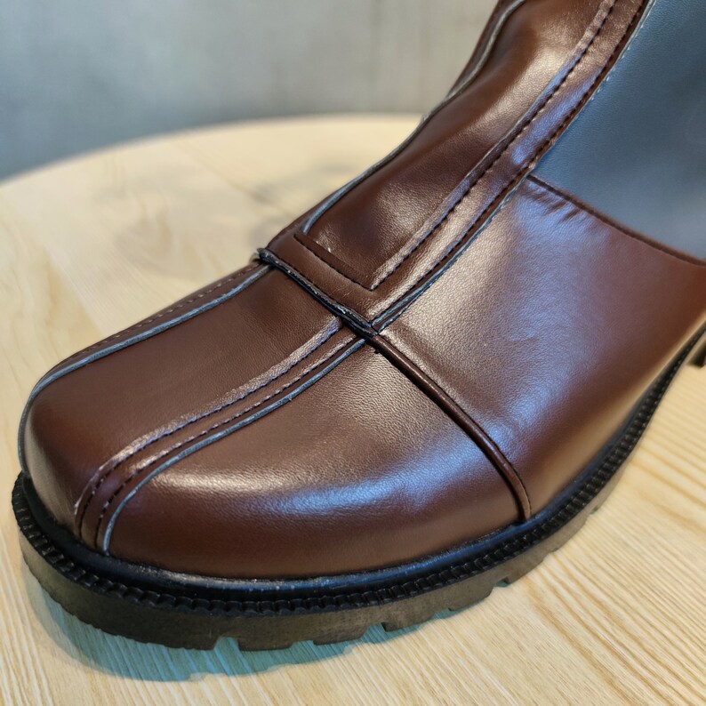 May include: Close-up of the toe of a brown leather boot with a black rubber sole. The boot has a seam down the center of the toe and a contrasting brown leather panel on the side.