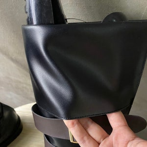K&ouml;nnte beinhalten: Schwarze Lederstiefel mit braunen Riemen und goldenen Schnallen. Die Stiefel haben einen umklappbaren Schaft mit einer Klappe, die eingesteckt werden kann.