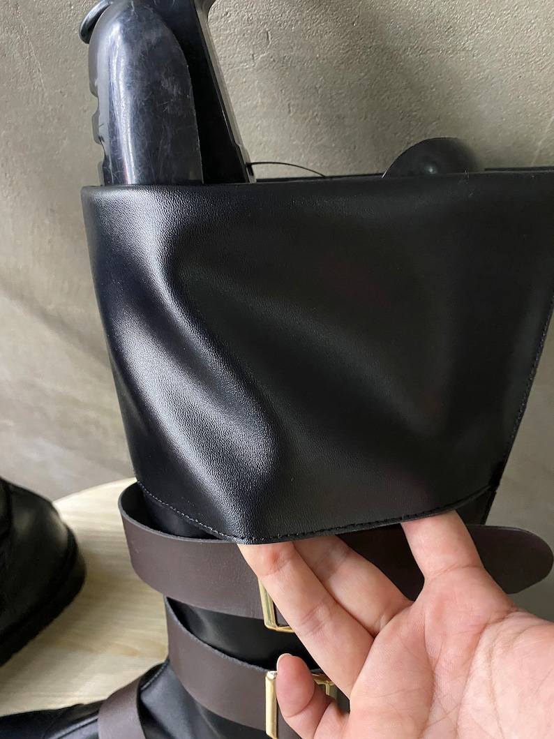 K&ouml;nnte beinhalten: Schwarze Lederstiefel mit braunen Riemen und goldenen Schnallen. Die Stiefel haben einen umklappbaren Schaft mit einer Klappe, die eingesteckt werden kann.