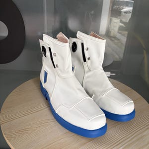 Puede incluir: Un par de botines blancos con acabado liso, suelas de goma azul y detalles rectangulares azules en los laterales. Los botines incluyen botones de presión de metal plateado, ribetes negros y un diseño de caña alta con silueta estructurada.