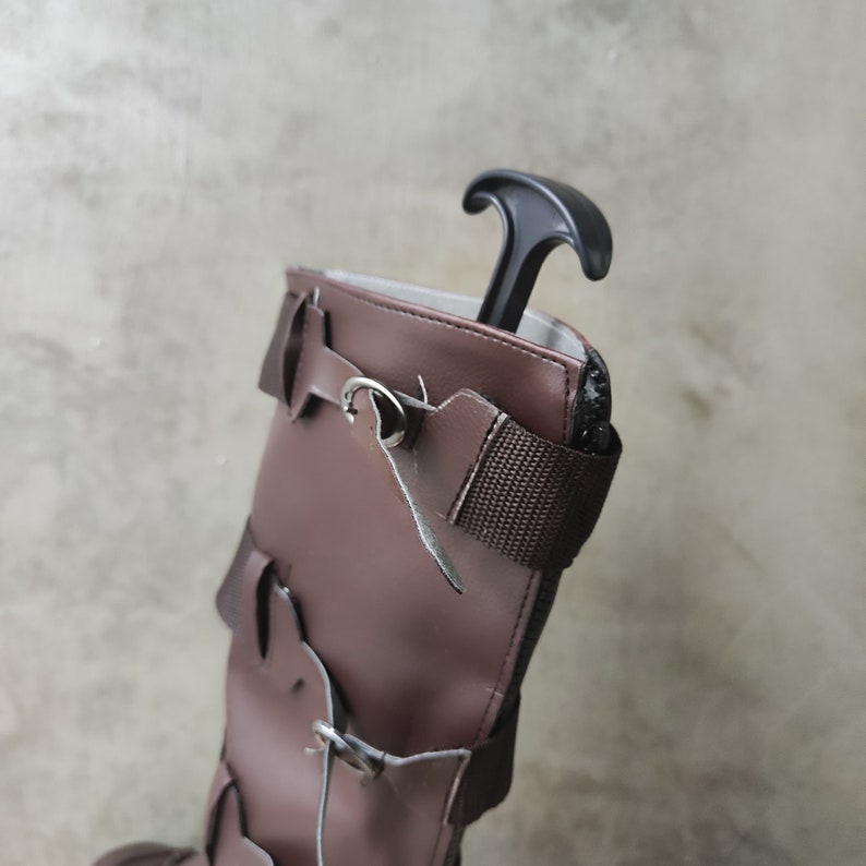 May include: A brown leather boot with buckles and straps. A black shoe horn is inserted into the top of the boot.