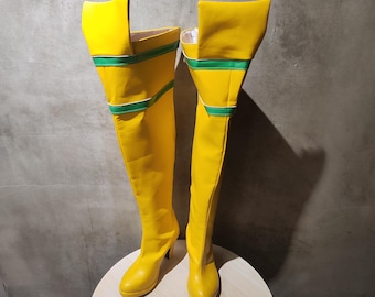 May include: A pair of bright yellow thigh-high boots with a high heel. The boots have a green stripe detail around the top and are made of a smooth material. The boots are displayed on a round, light-colored surface.
