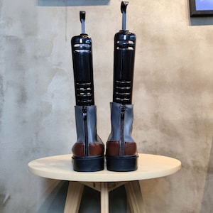 May include: A pair of gray and brown leather boots with black shoe trees inserted. The boots are on a wooden table.