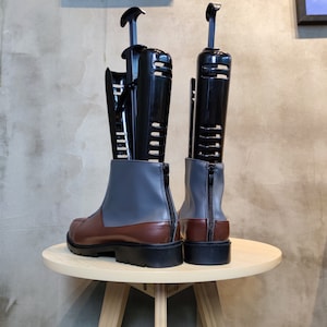 May include: A pair of brown and gray leather boots with black boot shapers inserted inside. The boots are on a wooden table.