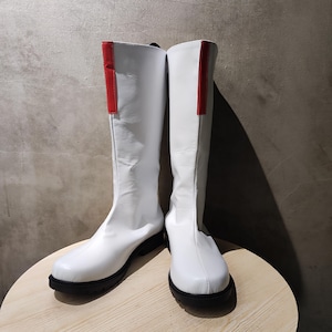 Könnte beinhalten: Ein Paar weiße, kniehohe Stiefel mit roten Akzenten an den Seiten. Die Stiefel haben eine abgerundete Spitze und eine schwarze Sohle.