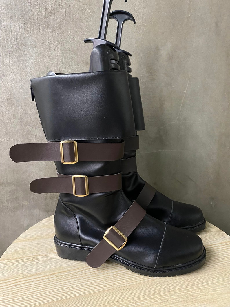 K&ouml;nnte beinhalten: Schwarze Lederstiefel mit braunen Riemen und goldenen Schnallen. Die Stiefel haben einen Rei&szlig;verschluss an der Seite und eine dicke Sohle.