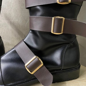 K&ouml;nnte beinhalten: Schwarze Lederstiefel mit braunen Lederriemen und goldenen Schnallen. Die Stiefel haben einen dicken Absatz und eine abgerundete Spitze.