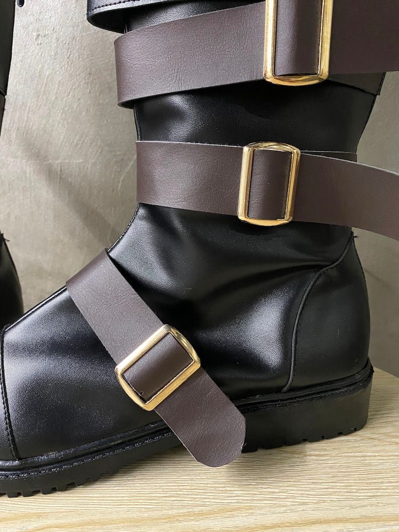K&ouml;nnte beinhalten: Schwarze Lederstiefel mit braunen Lederriemen und goldenen Schnallen. Die Stiefel haben einen dicken Absatz und eine abgerundete Spitze.
