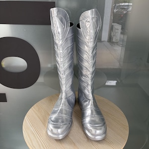 Puede incluir: Un par de botas plateadas hasta la rodilla. Las botas tienen un brillo metálico y presentan un diseño triangular en la parte superior. Las botas se exhiben sobre una mesa redonda de color marrón claro.