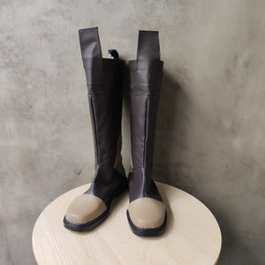 Puede incluir: Un par de botas de cuero marrón con detalles en cuero beige. Las botas tienen una punta puntiaguda y un eje alto.