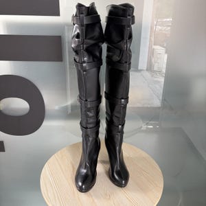 May include: A pair of black knee-high boots with multiple buckle straps. The boots have a pointed toe and a high heel. The upper portion of the boots is ruched, adding texture. The boots are displayed on a light-colored, round surface.