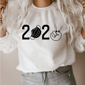 May include: White sweatshirt with black text and graphics that reads "2022" with a face mask and a hand making a peace sign with two syringes.