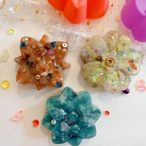 Op de afbeelding: Drie bloemvormige harsornamenten met ingebedde glitters, kleine kraaltjes en metalen accenten. Eén ornament is amberkleurig, één is iriserend wit met gouden spikkels en één is diepblauwgroen met een glinsterende afwerking vol glitters.
