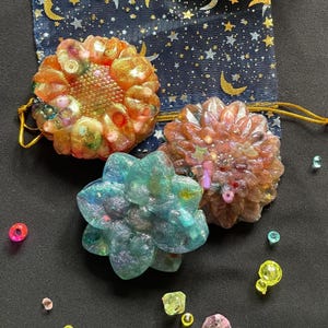 May include: Three resin flower-shaped objects in shades of orange, teal, and pink. The objects are embellished with glitter and small decorative elements. A navy blue drawstring bag with a gold moon and star pattern is in the background.