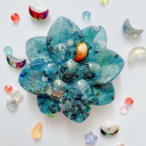 May include: A teal flower-shaped resin ornament embedded with glitter, small dark beads, and a single orange crescent moon charm. The piece is surrounded by various loose decorative beads and small crescent moon charms in assorted colors and finishes.
