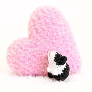 Pode incluir: Uma pequena figura de porquinho-da-&iacute;ndia de croch&ecirc; preto e branco em p&eacute; na frente de uma grande almofada em forma de cora&ccedil;&atilde;o rosa.