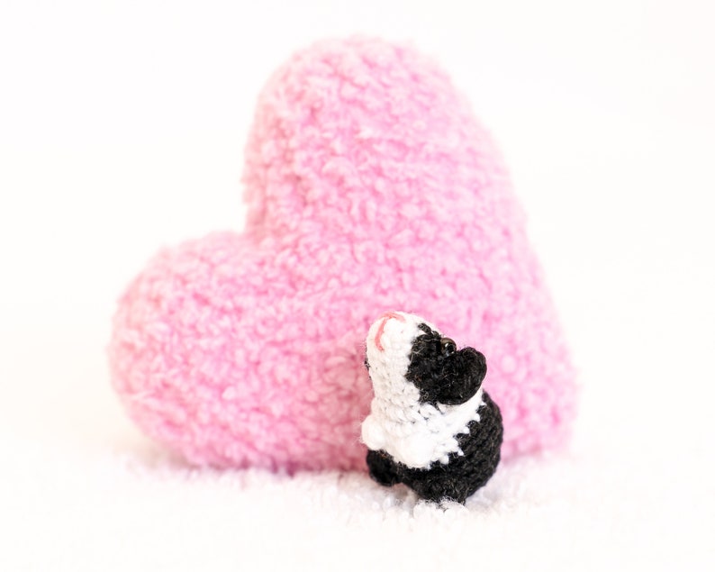 Pode incluir: Uma pequena figura de porquinho-da-&iacute;ndia de croch&ecirc; preto e branco em p&eacute; na frente de uma grande almofada em forma de cora&ccedil;&atilde;o rosa.
