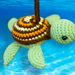 Puede incluir: Un juguete de tortuga marina de crochet con una concha marr&oacute;n, amarilla y blanca y un cuerpo verde. La tortuga est&aacute; colgada de una cuerda y parece estar bajo el agua.