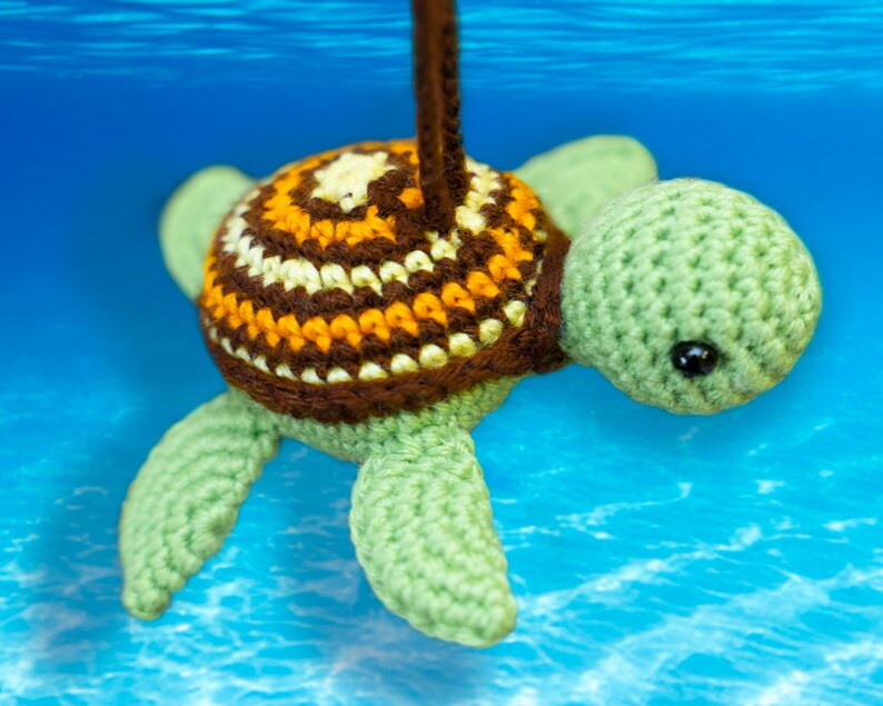 Puede incluir: Un juguete de tortuga marina de crochet con una concha marr&oacute;n, amarilla y blanca y un cuerpo verde. La tortuga est&aacute; colgada de una cuerda y parece estar bajo el agua.
