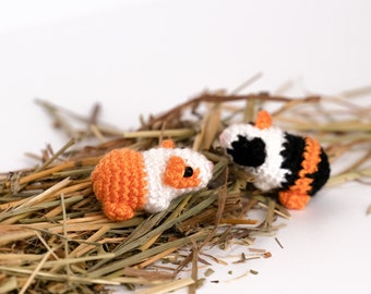 May include: Two crocheted guinea pig toys, one orange and white, the other black and white, are nestled in a bed of dried grass.
