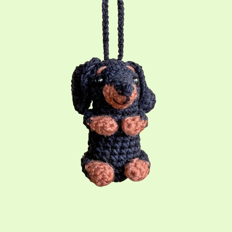 May include: A handmade crochet dachshund dog ornament. The dog is black with brown paws, ears, and snout. It has a black hanging loop. The dog is standing upright with its paws crossed.