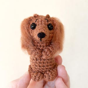 May include: A handmade, brown crochet dachshund dog with black eyes and a black nose. The dog has long, fluffy ears and is holding its paws up. The crochet dog is a charming, whimsical creation.