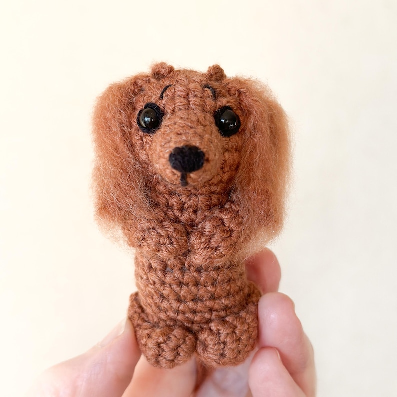 May include: A handmade, brown crochet dachshund dog with black eyes and a black nose. The dog has long, fluffy ears and is holding its paws up. The crochet dog is a charming, whimsical creation.