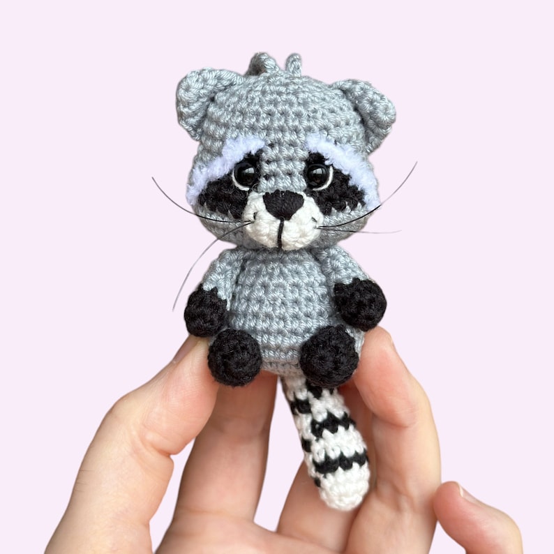 May include: A crocheted raccoon plush toy with a gray body, black eyes, a white nose, and a black and white striped tail.