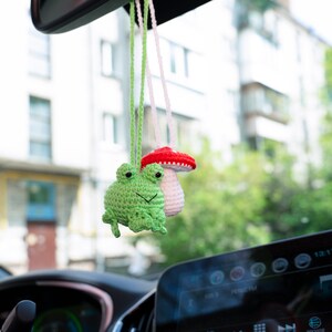 Puede incluir: Una rana de crochet verde y un hongo de crochet blanco con la parte superior roja cuelgan del espejo retrovisor de un coche.