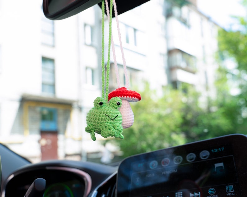 Puede incluir: Una rana de crochet verde y un hongo de crochet blanco con la parte superior roja cuelgan del espejo retrovisor de un coche.