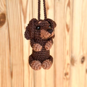 May include: A crocheted dachshund dog ornament in shades of brown, with a dark brown body and lighter brown paws and snout. The dog has black eyes and is suspended by a matching brown cord, perfect for hanging. The background is a light brown wooden surface.