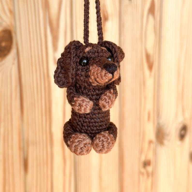 May include: A crocheted dachshund dog ornament in shades of brown, with a dark brown body and lighter brown paws and snout. The dog has black eyes and is suspended by a matching brown cord, perfect for hanging. The background is a light brown wooden surface.