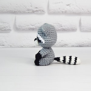 May include: A crocheted raccoon plush toy with a gray body, black eyes, white nose, and a black and white striped tail.