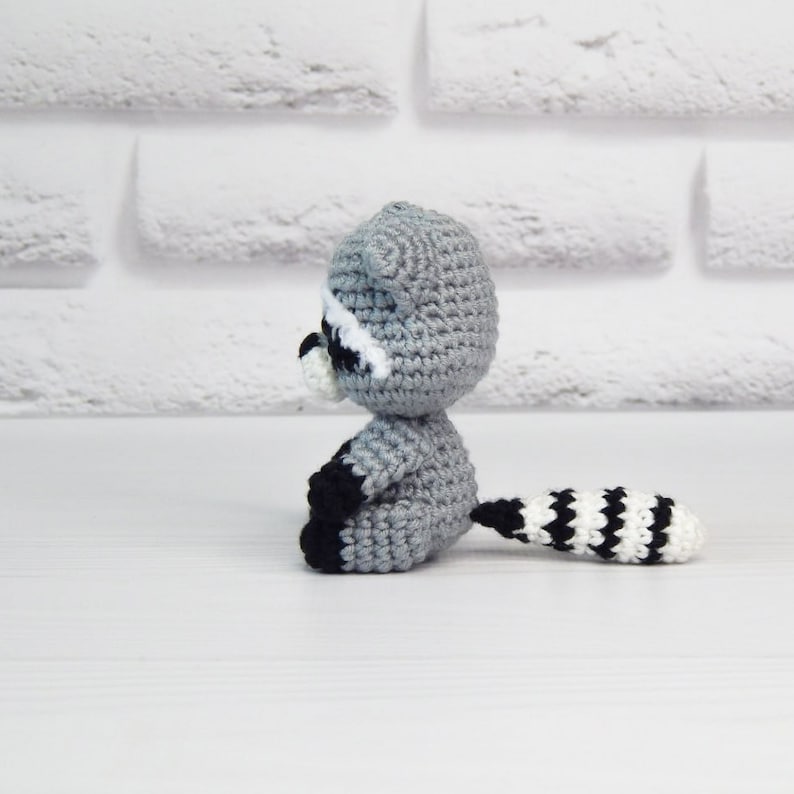 May include: A crocheted raccoon plush toy with a gray body, black eyes, white nose, and a black and white striped tail.