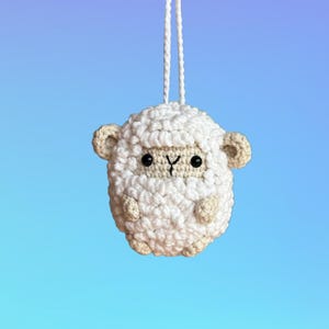 Puede incluir: Un adorno de oveja de crochet blanco con ojos negros y una peque&ntilde;a nariz rosa. La oveja est&aacute; colgada de una cuerda blanca.