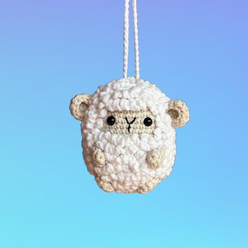 Puede incluir: Un adorno de oveja de crochet blanco con ojos negros y una peque&ntilde;a nariz rosa. La oveja est&aacute; colgada de una cuerda blanca.
