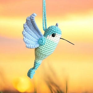 May include: A crocheted hummingbird ornament with blue and green wings, a white belly, and a black beak. The ornament is hanging from a string.