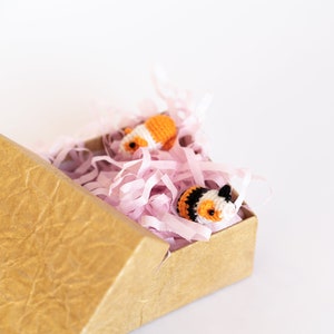 Pode incluir: Duas pequenas figuras de animais de croch&ecirc;, uma laranja e branca, a outra preta, branca e laranja, est&atilde;o aninhadas em papel crepom rosa dentro de uma caixa de papel&atilde;o marrom.