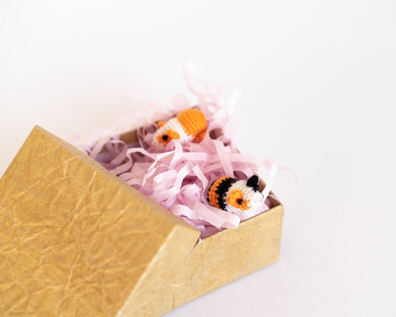 Pode incluir: Duas pequenas figuras de animais de croch&ecirc;, uma laranja e branca, a outra preta, branca e laranja, est&atilde;o aninhadas em papel crepom rosa dentro de uma caixa de papel&atilde;o marrom.