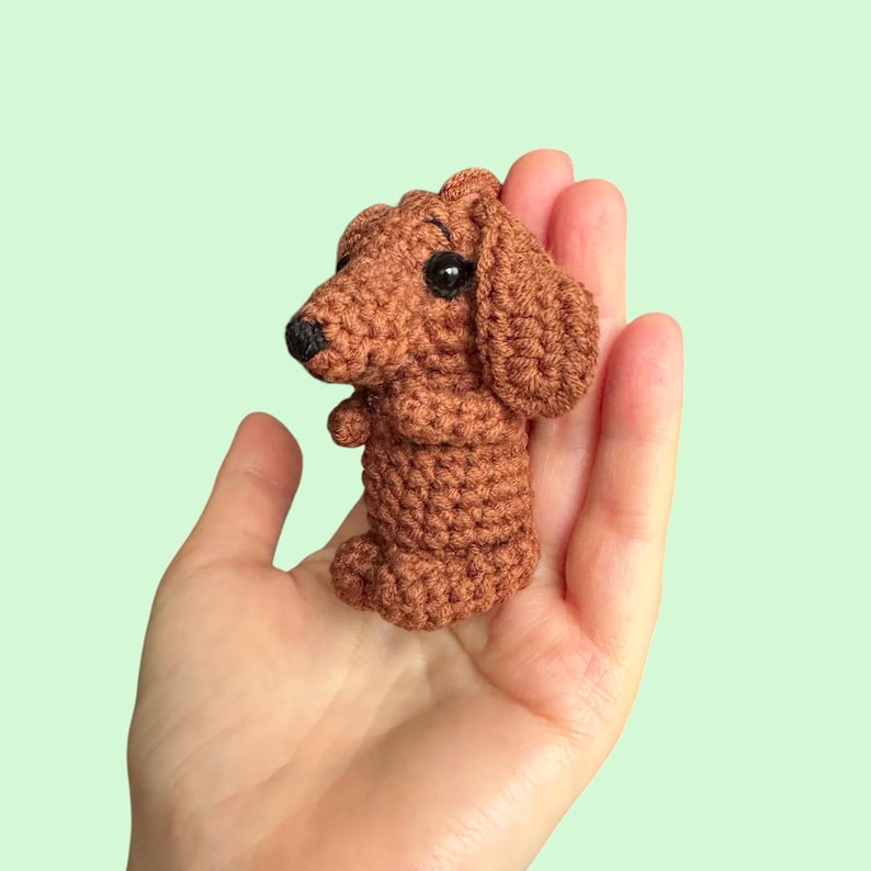 May include: A crocheted brown Dachshund dog figurine with black eyes and nose. The handmade dog has floppy ears and is held in a hand against a light green background. The dog is approximately 3 inches tall.
