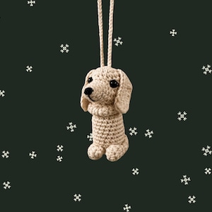May include: A crocheted dog ornament in a light beige color, with black eyes and a black nose. The dog has floppy ears and is suspended by a matching beige cord. The background is a dark green with white snowflake accents.