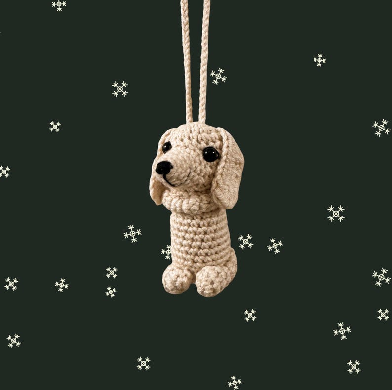 May include: A crocheted dog ornament in a light beige color, with black eyes and a black nose. The dog has floppy ears and is suspended by a matching beige cord. The background is a dark green with white snowflake accents.