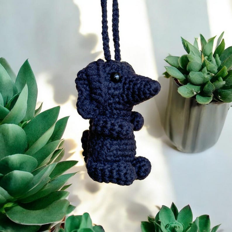 May include: A navy blue crocheted dachshund dog ornament with black bead eyes and a hanging loop. The dog is positioned in front of green succulents in a gray pot and other green plants. The dog is approximately 4 inches tall.
